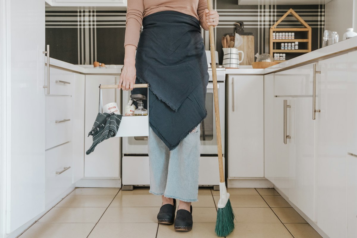 Housekeeper with a broom and cleaning caddy in a bright white kitchen